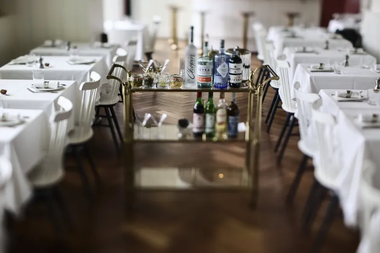 Bar cart with assorted liquor bottles and glasses in an elegant restaurant dining area with white tablecloths.