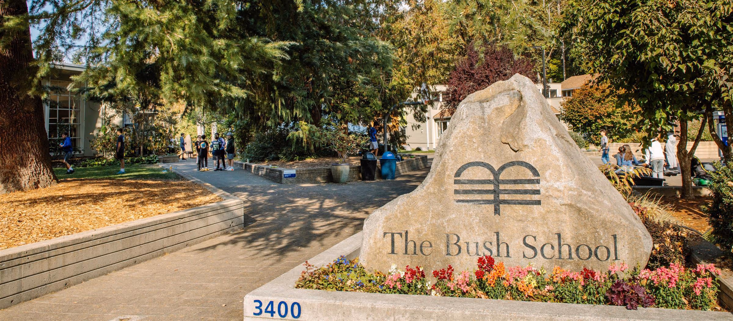 Stone sign for The Bush School surrounded by flowers with students and trees in the background.