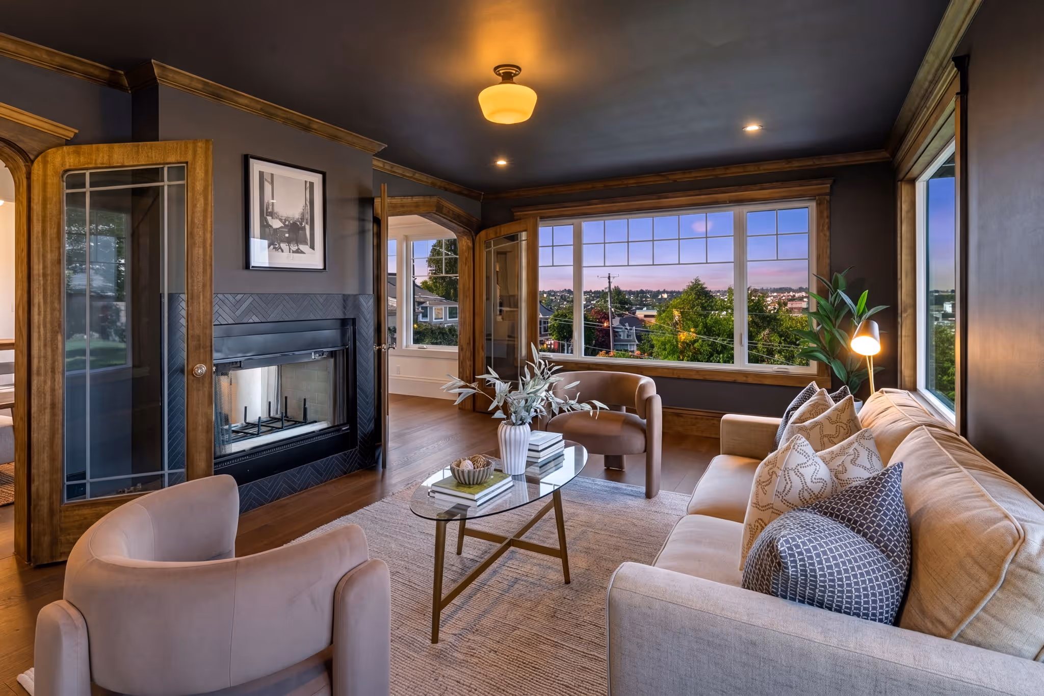 Modern living room with beige sofa, glass coffee table, armchairs, and large windows showing a city view at dusk.