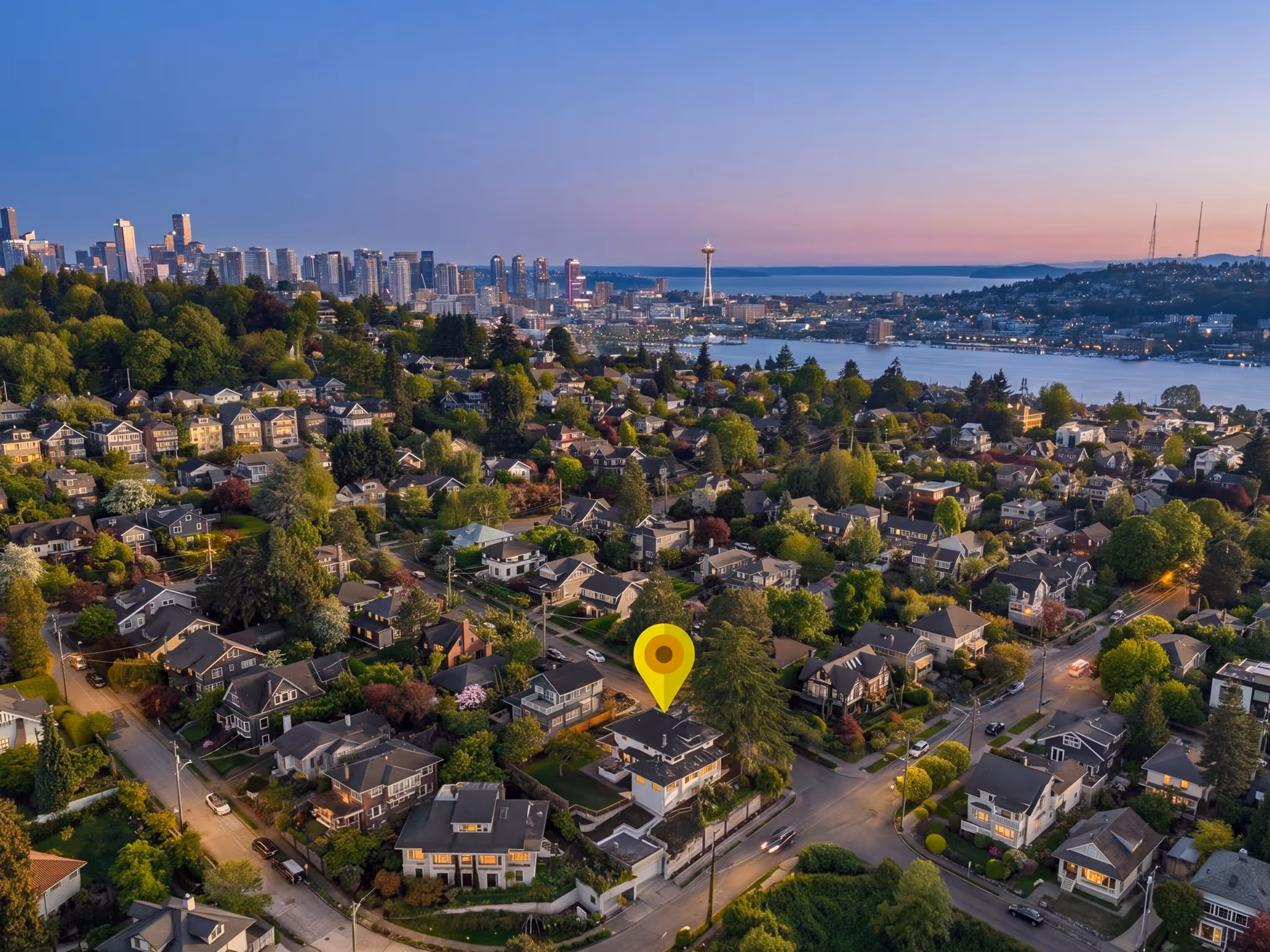 Aerial view of a Seattle neighborhood with a yellow location pin and the city skyline in the background at sunset.