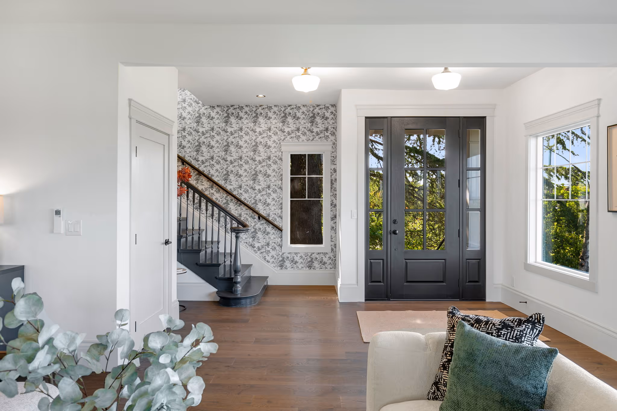 Bright home entry with black front doors, patterned wallpaper on stair wall, hardwood floors, and modern furniture.