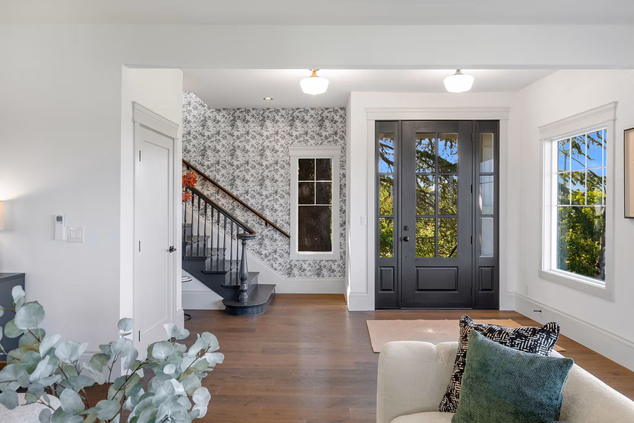 Bright entryway with dark wooden front door, floral wallpapered staircase, light hardwood floors, and large windows.