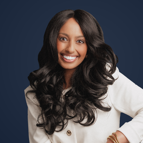 Smiling woman with long wavy black hair wearing a light coat against a dark blue background.