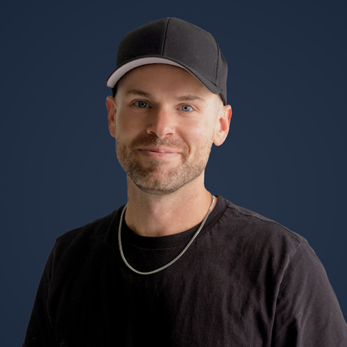 Man with short beard wearing black cap, black shirt, and silver chain necklace against a dark background.