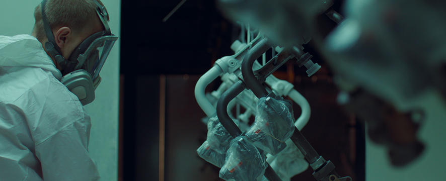 Person in a protective suit and mask spray paints objects hanging in a dimly lit area.