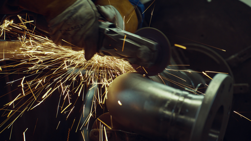 Worker using a grinder on metal pipe, creating bright sparks flying in multiple directions.