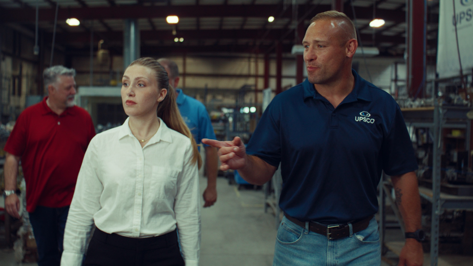 Man in UPSCO shirt gestures and talks while walking with three coworkers in a warehouse.