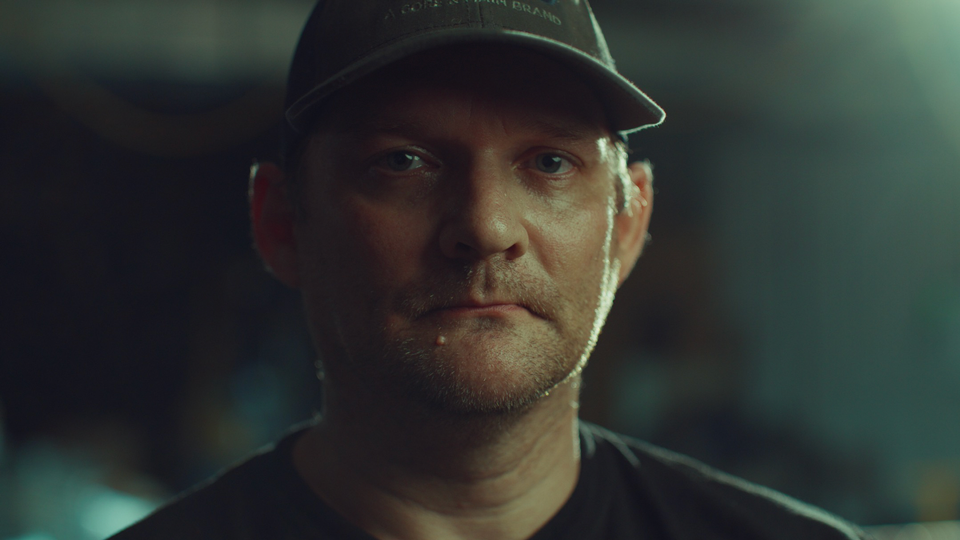 Close-up of a serious man wearing a dark cap and shirt against a blurred background.