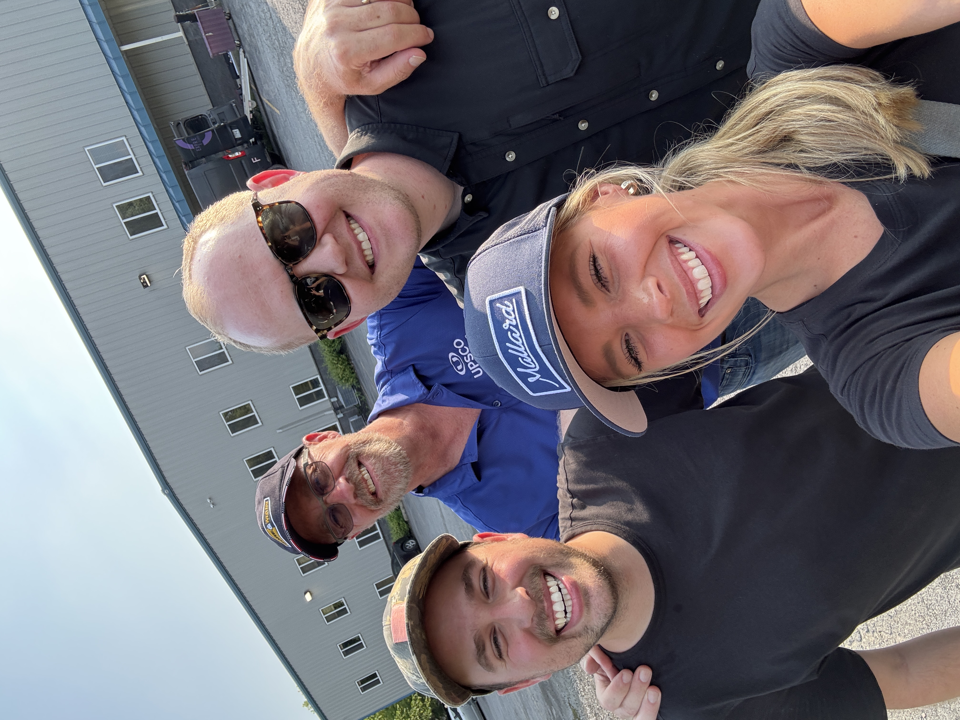 Four people smiling outdoors in front of a large industrial building on a sunny day.