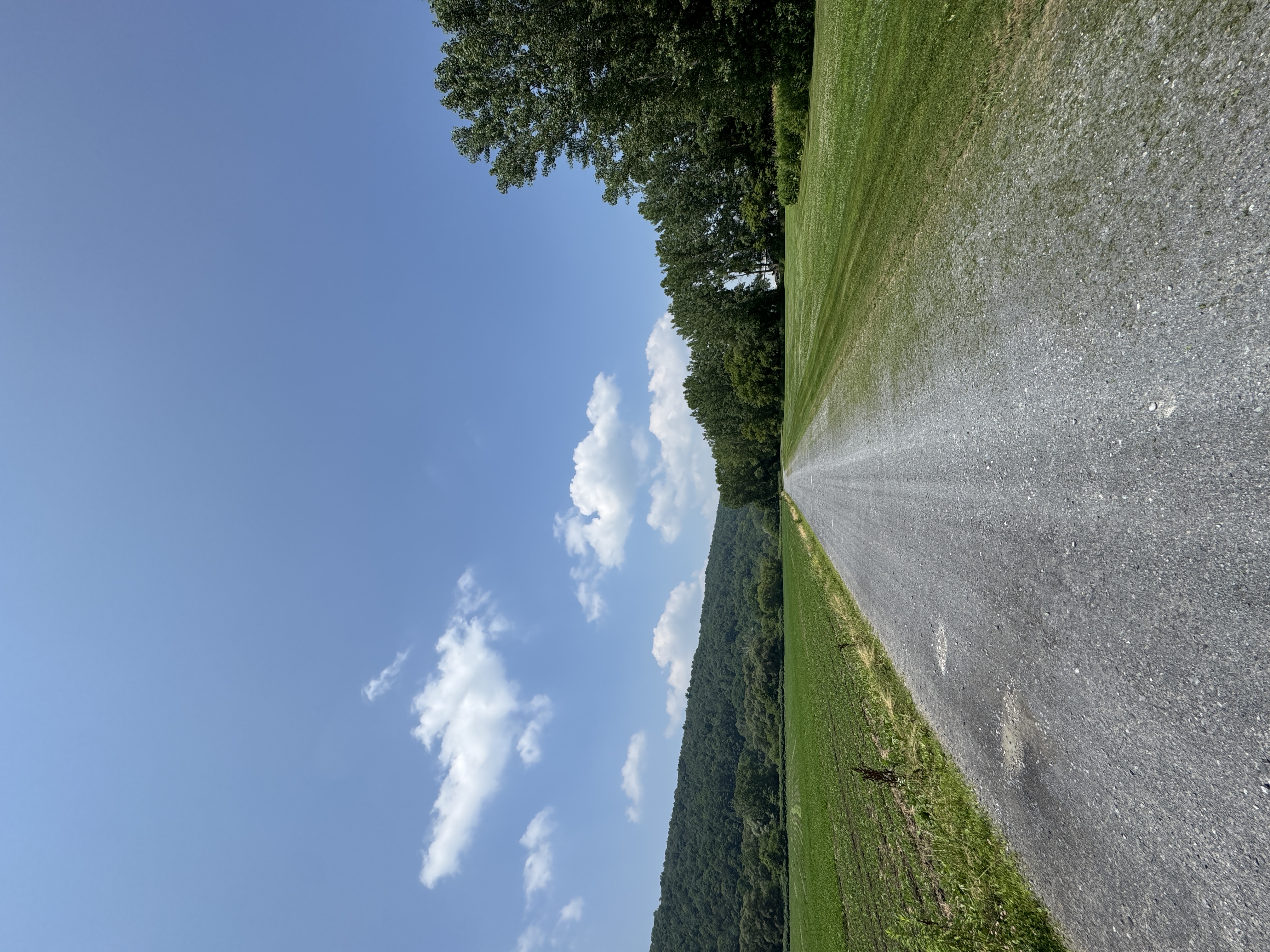 Gravel path runs between green fields under a blue sky with scattered clouds and distant hills.