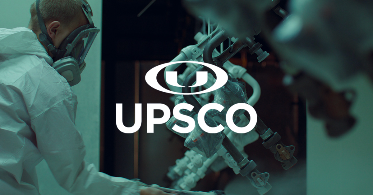 Person in protective suit and mask working near machinery pipes in a dark industrial setting with UPSCO logo.