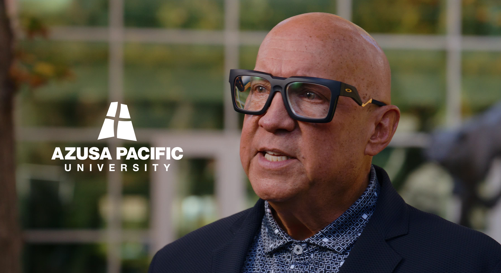 Man wearing large black glasses and a patterned shirt with Azusa Pacific University logo beside him.