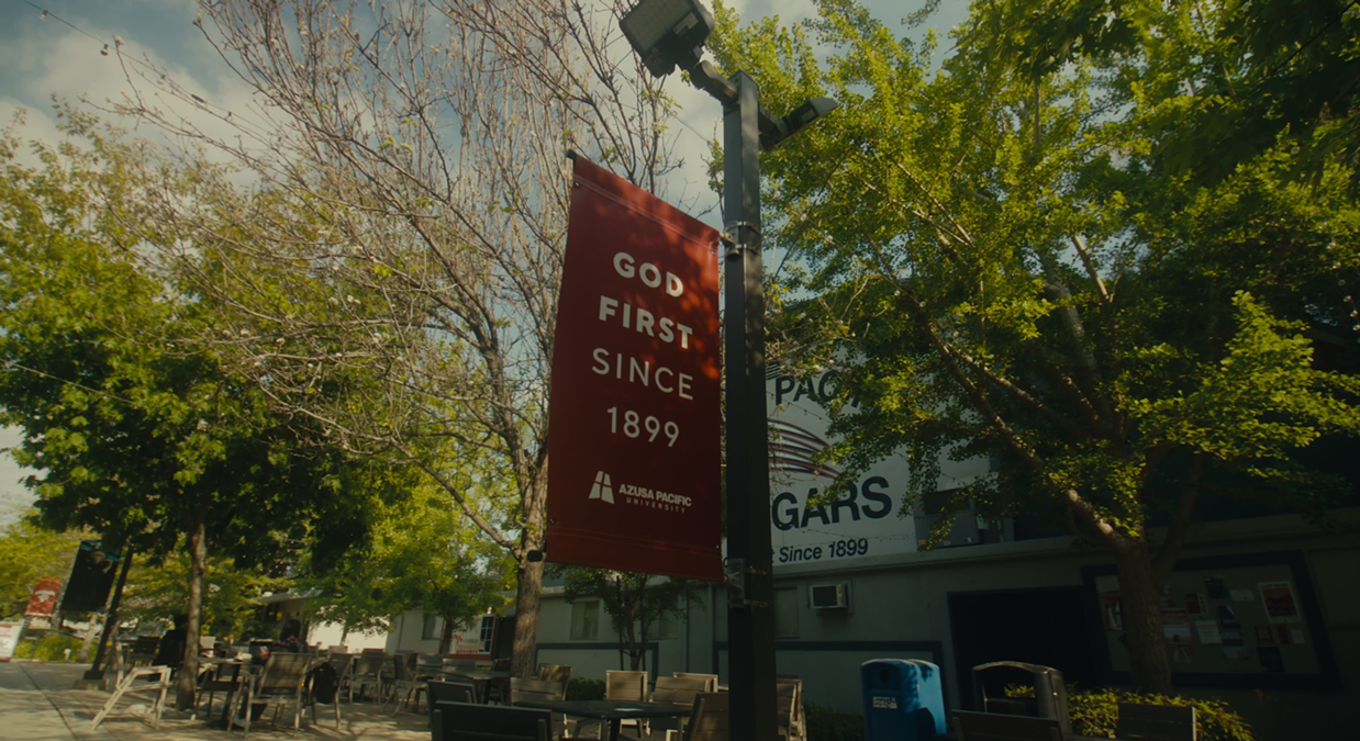 Red banner on a lamp post reads God First Since 1899 at Azusa Pacific University campus.