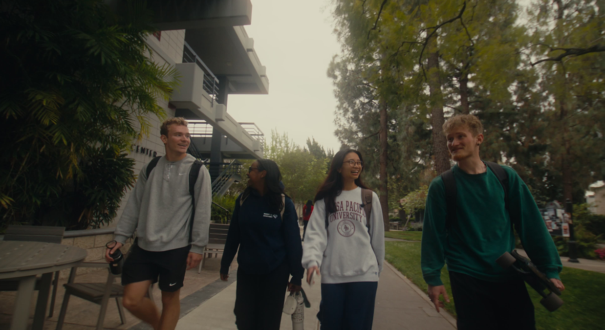 Four college students walking and smiling on a campus sidewalk surrounded by trees and buildings.