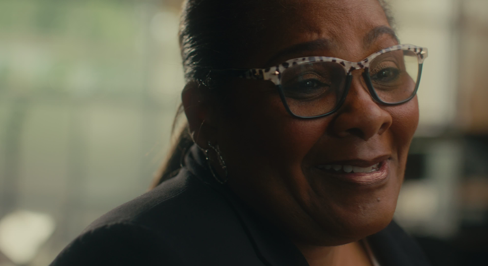 Close-up of a smiling woman with glasses and hoop earrings indoors.