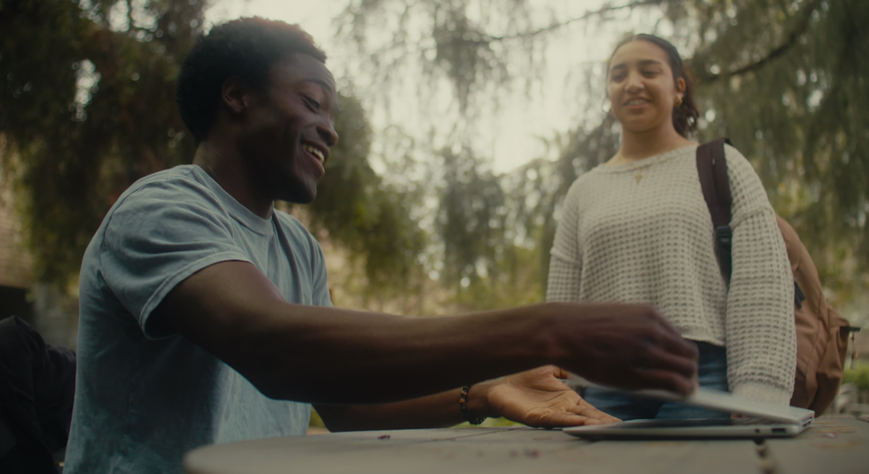 Smiling man sitting at outdoor table handing tablet to standing woman with backpack.