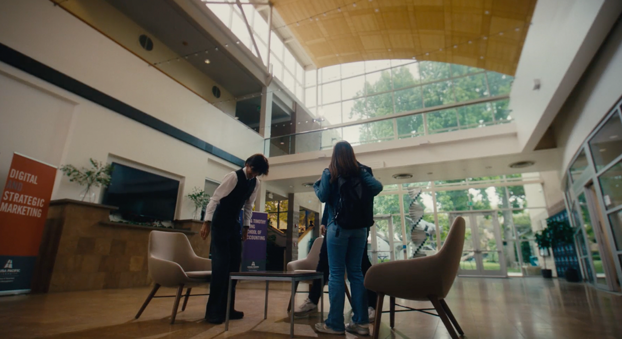 Three people talking in a modern lobby with large windows, chairs, and marketing banners.