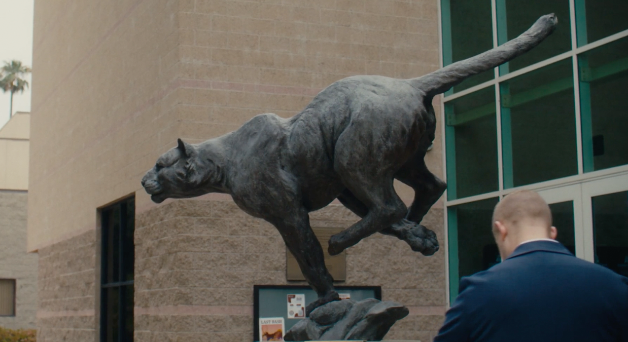 Bronze statue of a cougar mid-leap outside a building with a man in a blue jacket nearby.