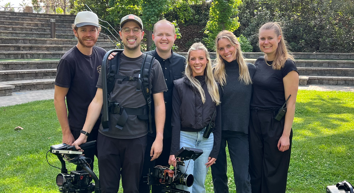 Group of six smiling people with video cameras standing outdoors on grass in front of stone steps.
