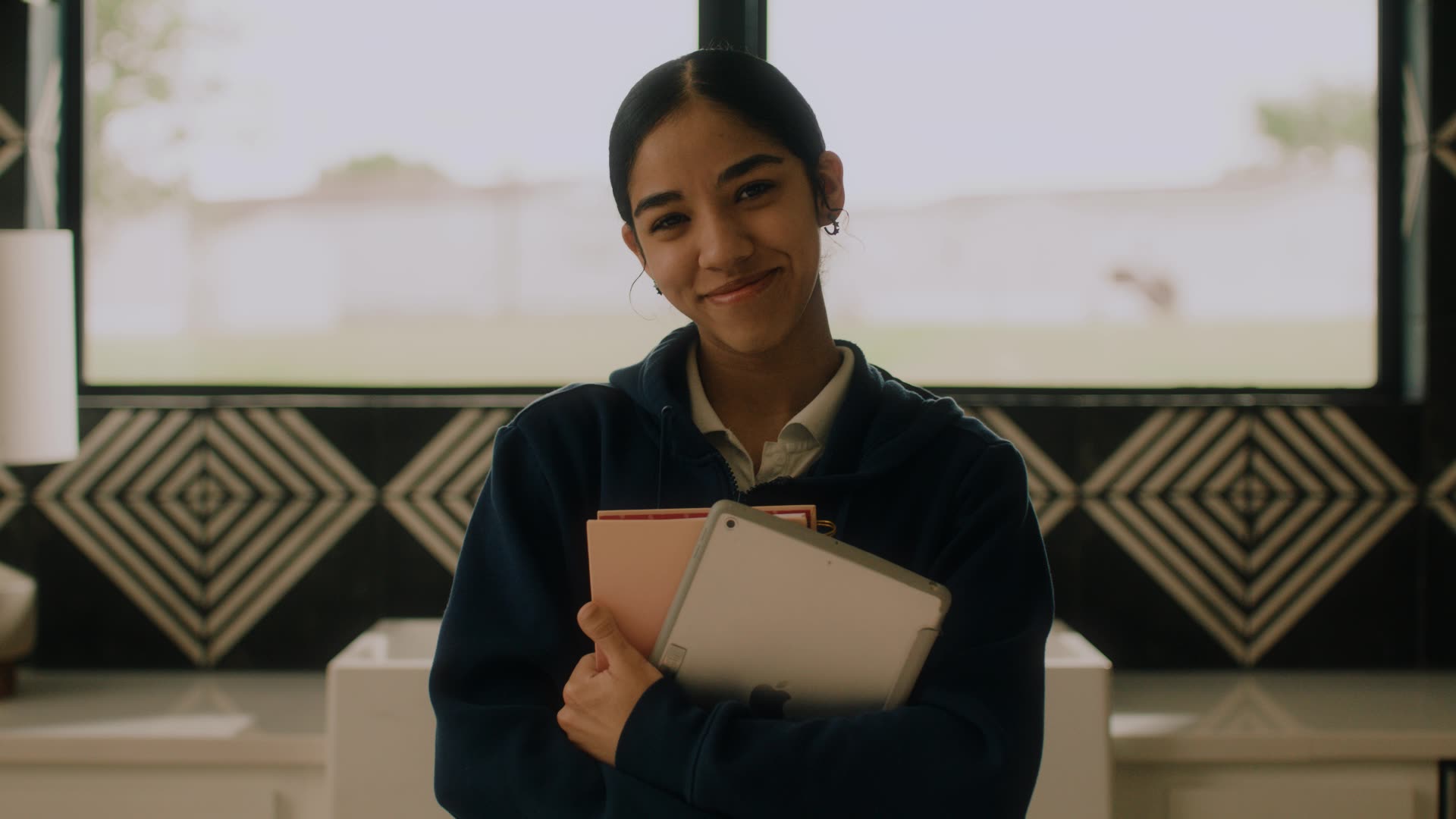 Young woman smiling and holding a tablet and notebooks indoors by a window with geometric decor.