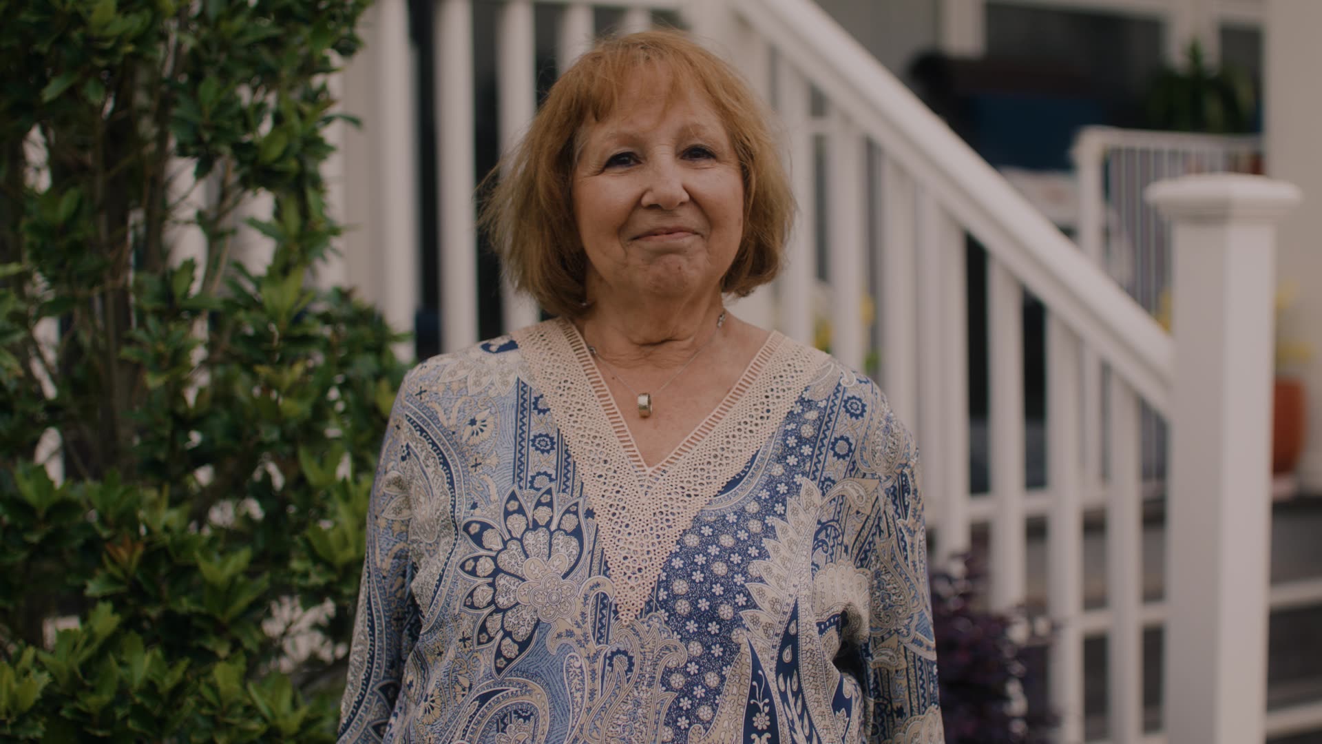 Smiling older woman with short brown hair wearing a patterned blue and beige top outside by stairs.