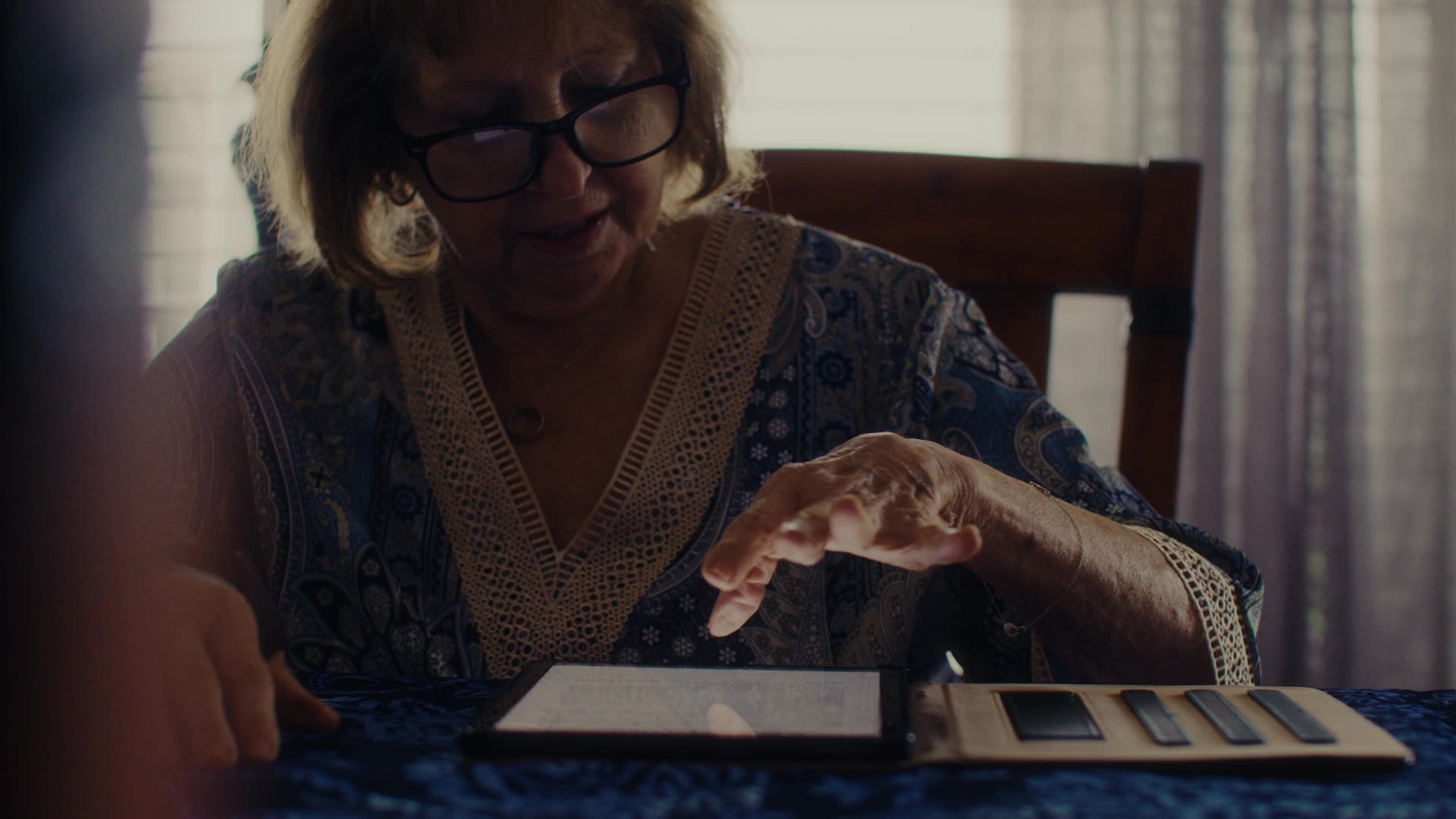 Older woman wearing glasses interacts with a tablet at a table with a case for the device nearby.