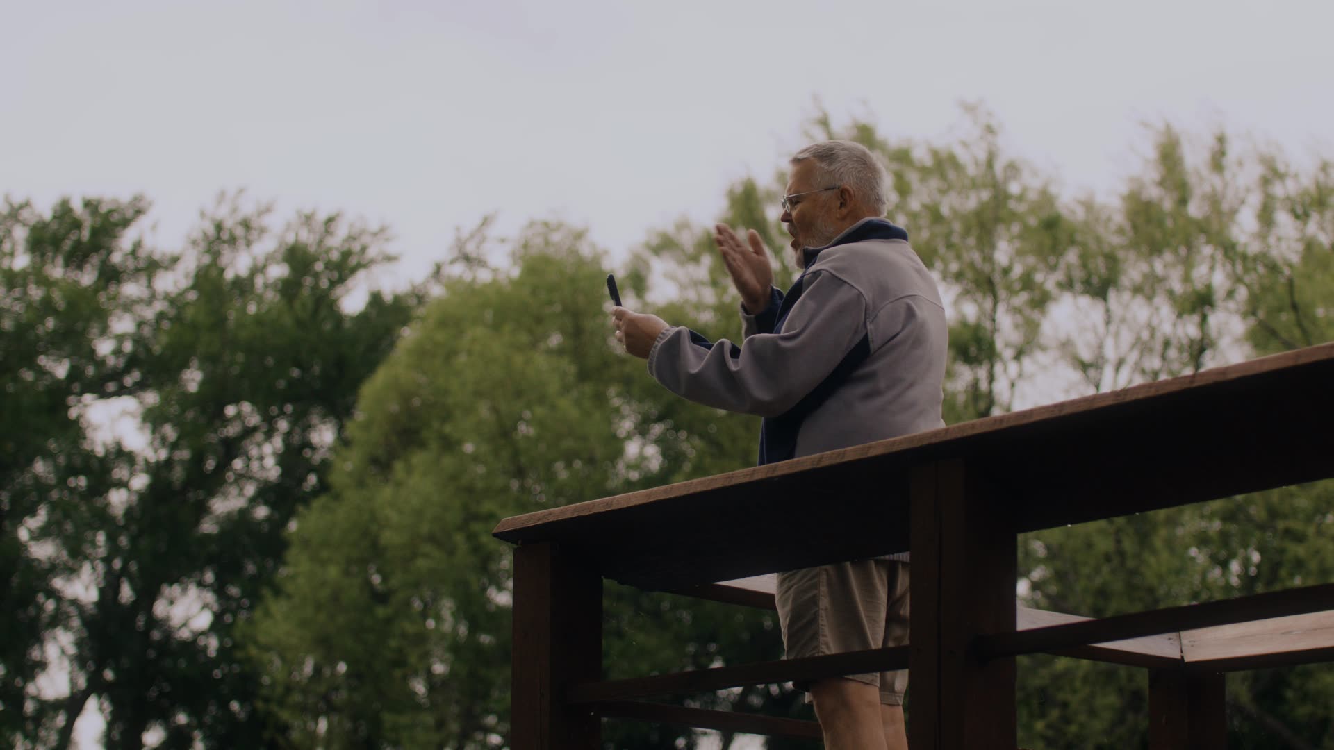 Older man standing on wooden deck outdoors, holding phone and gesturing with one hand.