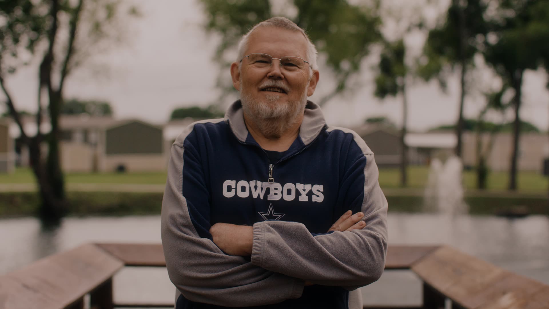 Smiling elderly man with glasses stands outdoors wearing a Cowboys sweatshirt with arms crossed.