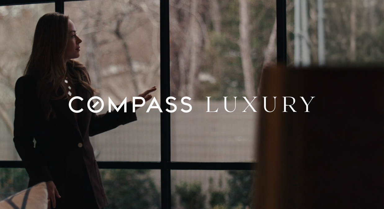 Woman in black coat standing by a large window with trees outside and Compass Luxury text overlay.