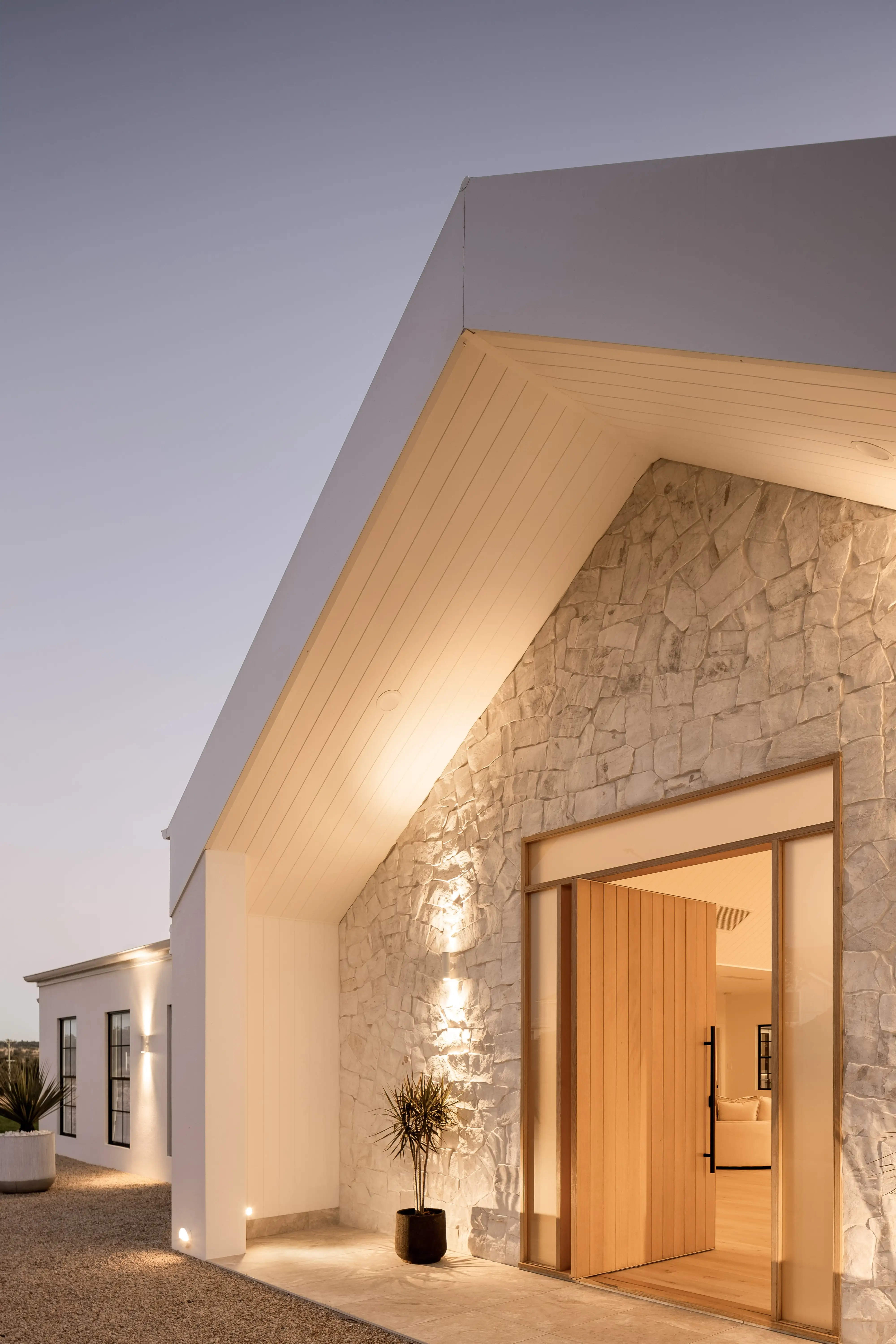 Modern farmhouse exterior from The Renew Company’s Callista Project featuring a stone façade, large wooden pivot door, and warm architectural lighting at dusk.