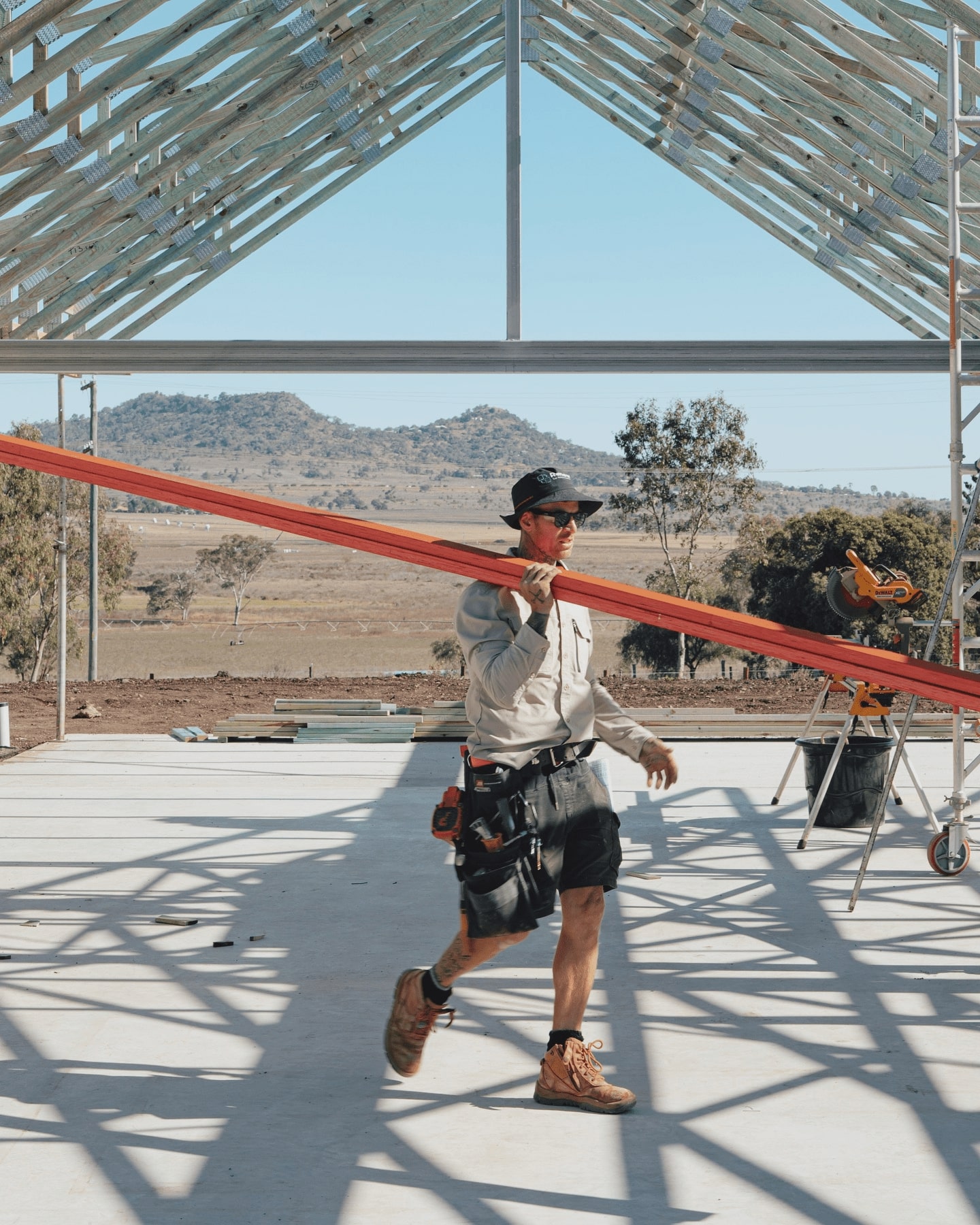 Construction worker from The Renew Company carrying a timber beam on a residential building site with metal roof trusses and scenic hills in the background.