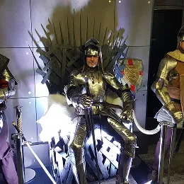 Une personne portant une armure de chevalier médiéval, assise sur un trône en métal rappelant le Trône de Fer.