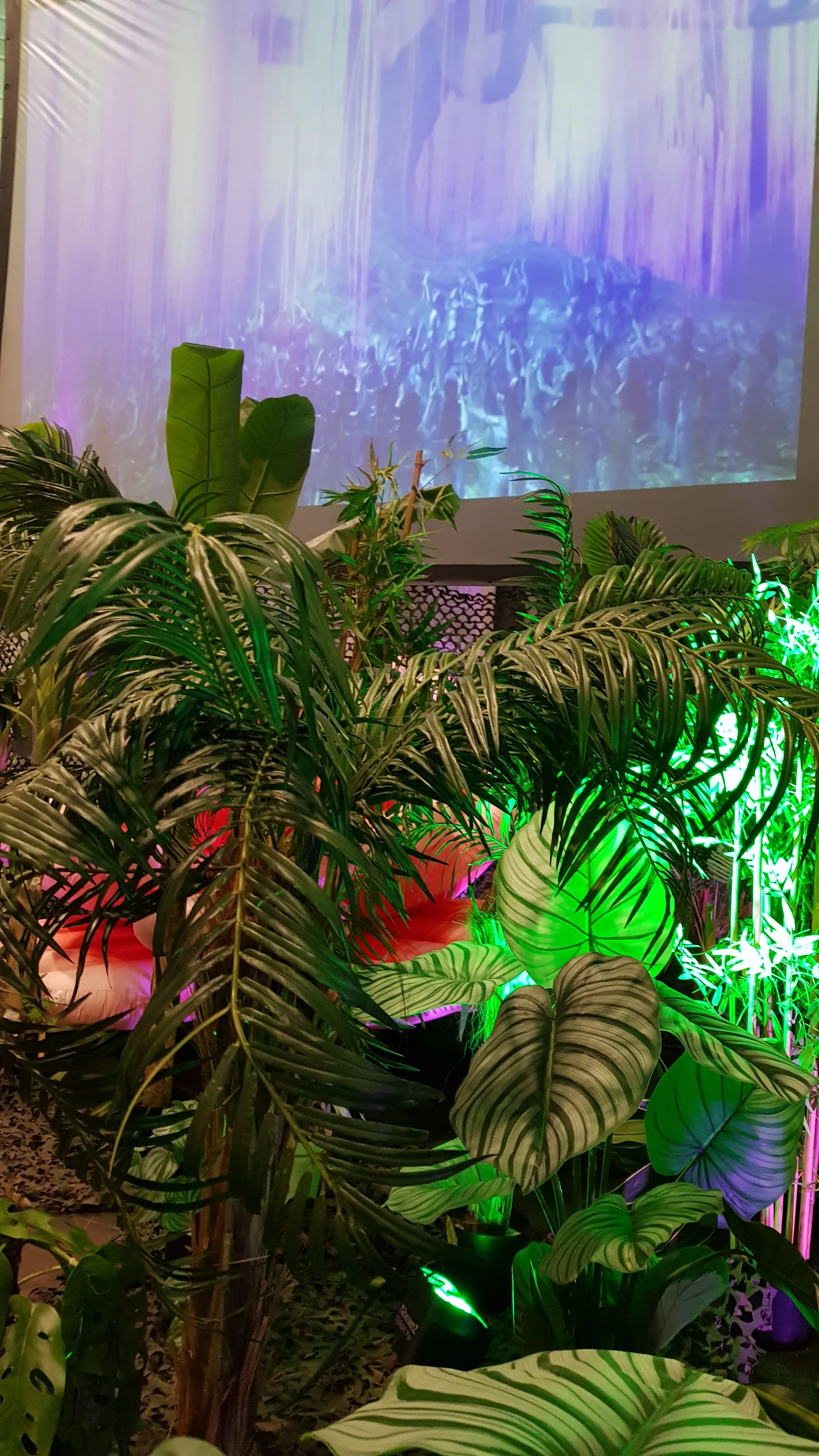 Décor d'événement simulant une cascade avec des palmiers et des plantes tropicales éclairés en vert et rouge.