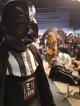 Une personne portant un costume complet et détaillé de Dark Vador, incluant le casque et la cape, se tenant debout lors d'un événement en intérieur.