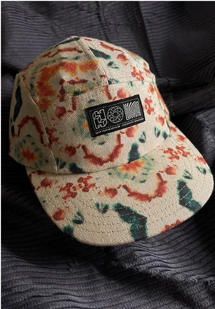 Beige fabric baseball cap with red, green, and orange abstract patterns and a black rectangular patch with white geometric designs on the front.