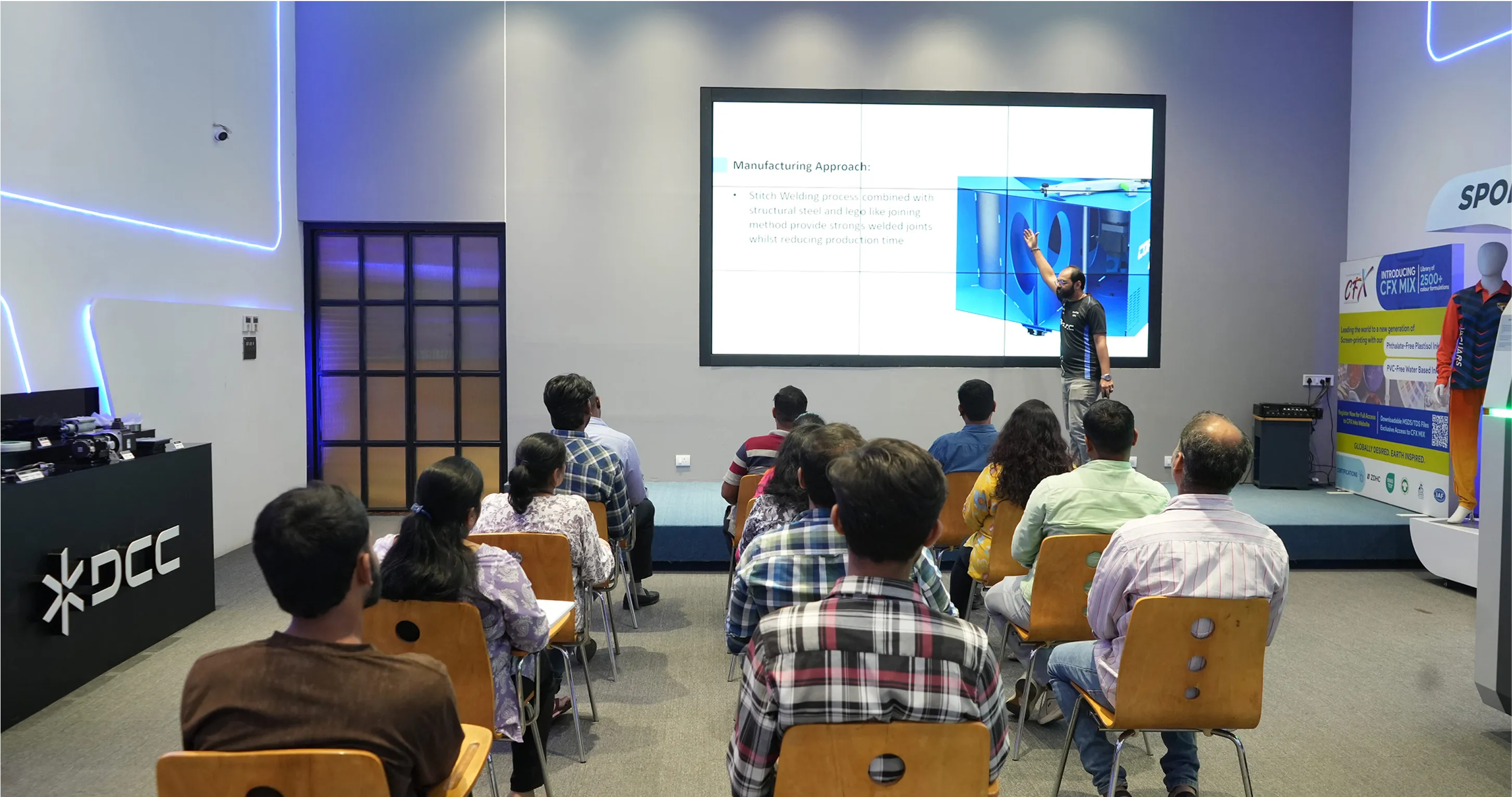 A man giving a presentation to an audience seated in rows, with a large screen displaying manufacturing process details behind him in a modern conference room.