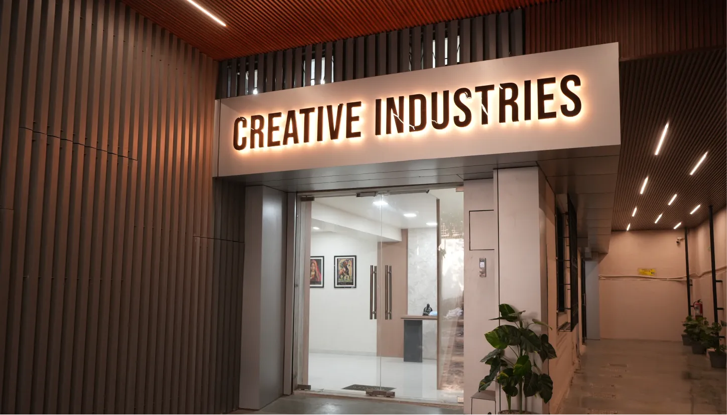 Entrance to a building with a glowing sign reading 'CREATIVE INDUSTRIES' above glass doors and potted plants nearby.