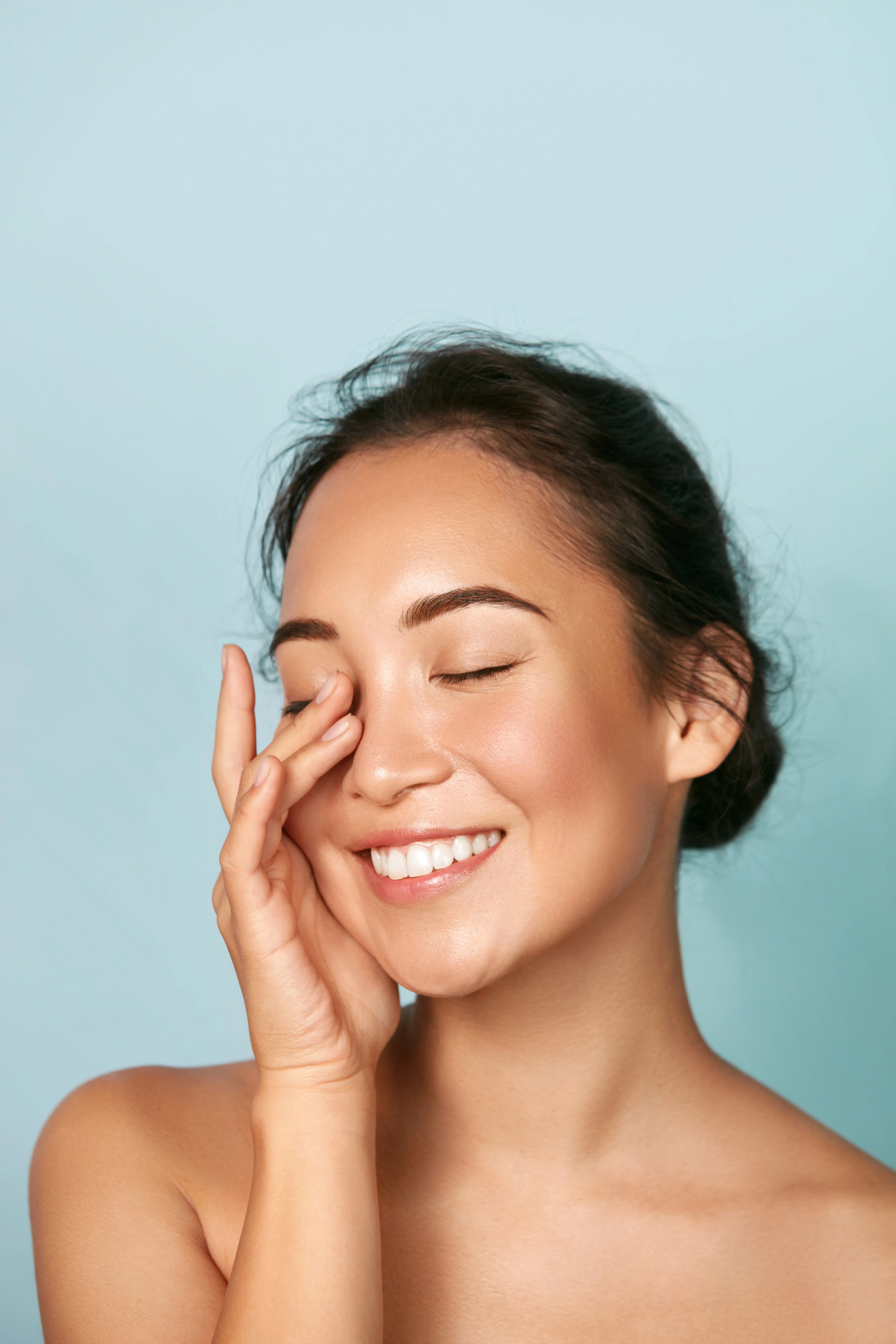 Smiling woman with clear skin gently touching her face with eyes closed against a light blue background.