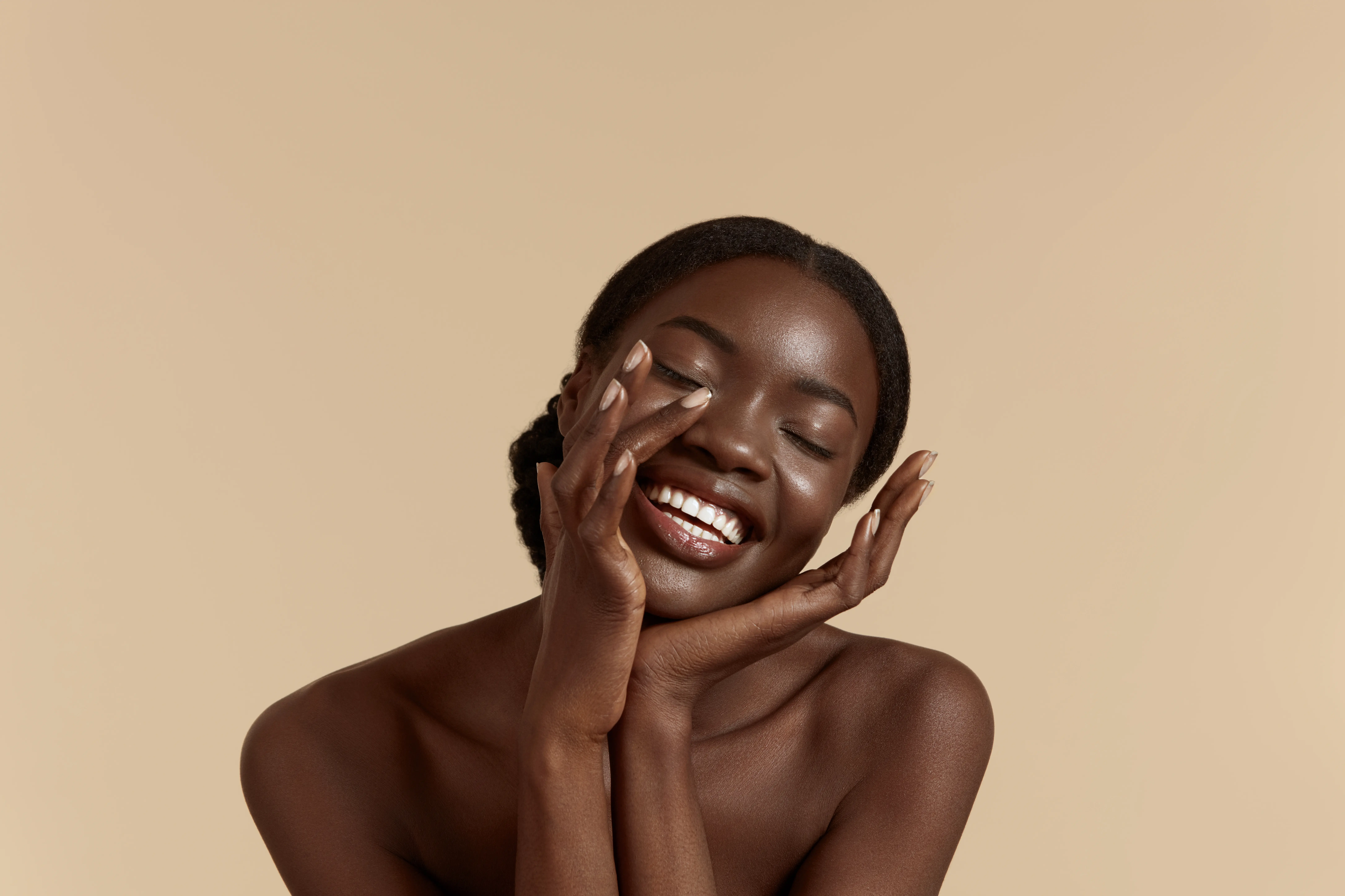 Woman with dark skin and hair pulled back smiling with eyes closed, hands gently touching her face against a beige background.