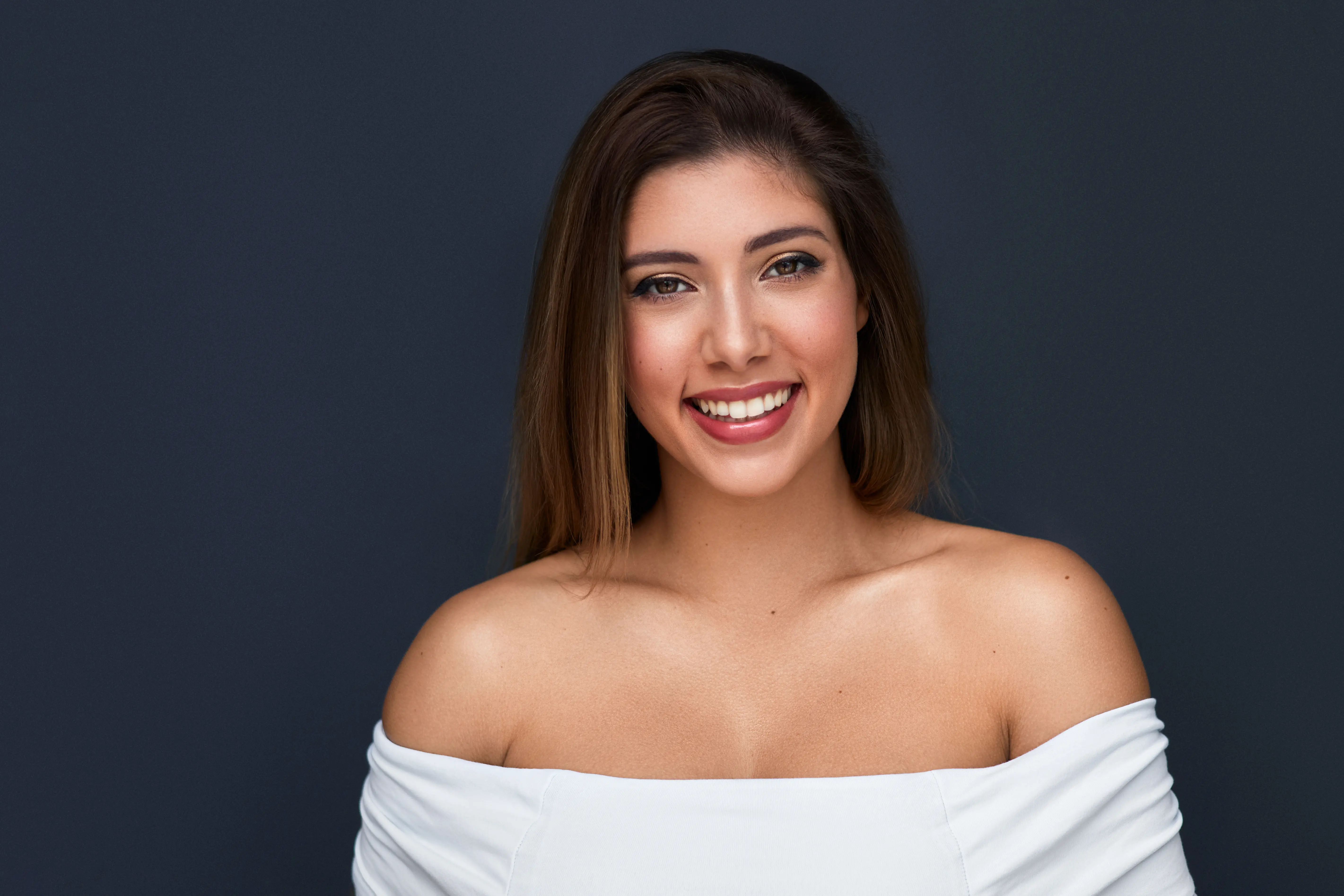 Smiling young woman with long brown hair wearing a white off-shoulder top against a dark background.