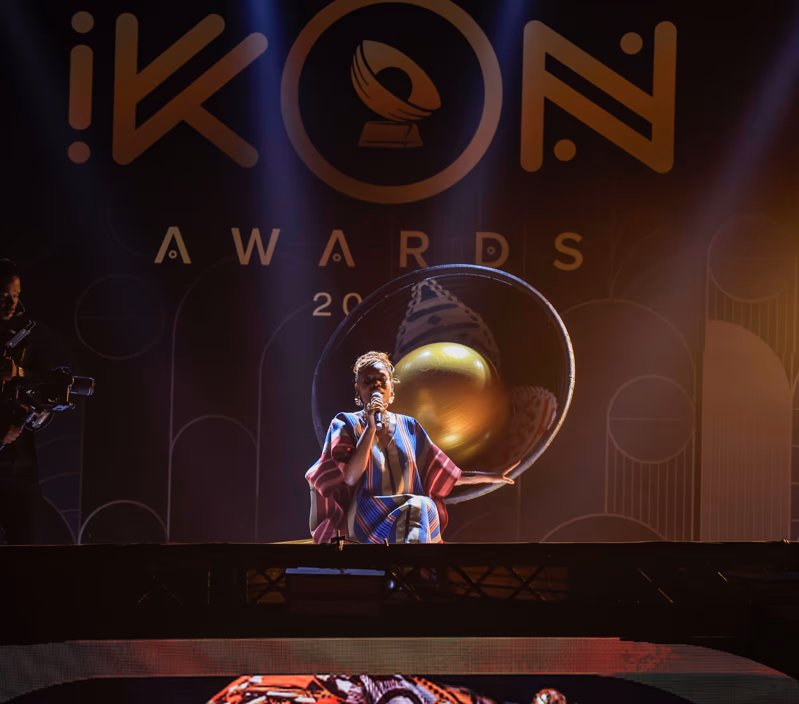 Woman in patterned dress sings on stage with a large golden award trophy behind her at iKON Awards.