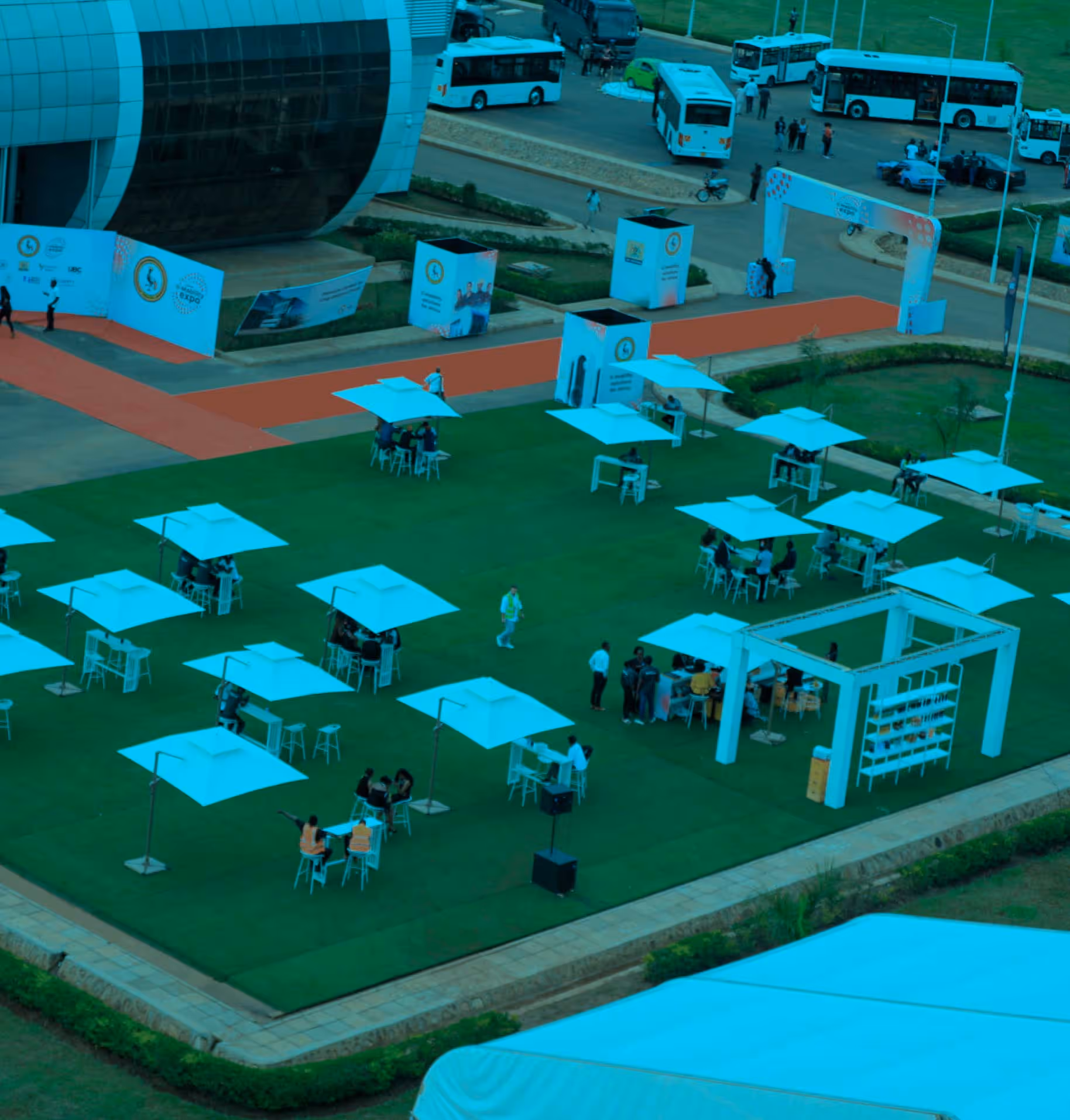 Outdoor event with people sitting under white umbrellas on green grass near a large modern building.