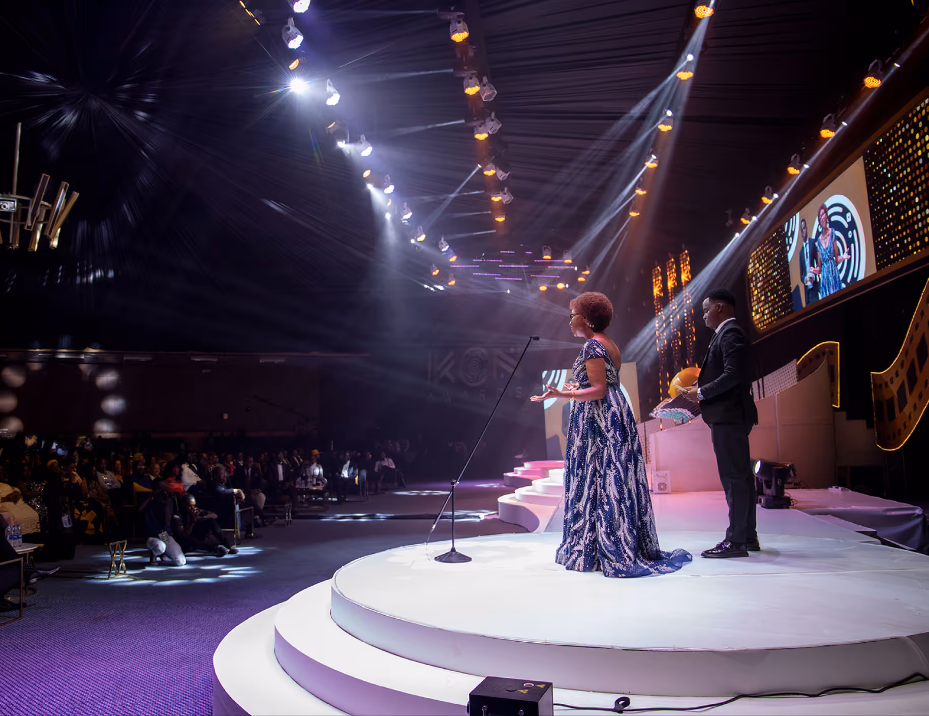 Woman in patterned gown and man in suit on stage speaking to audience under spotlight.