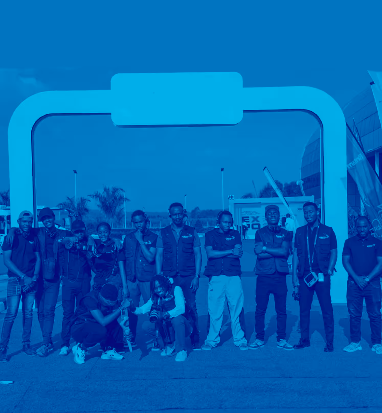 Group of people posing outdoors in front of a large archway, some holding cameras.