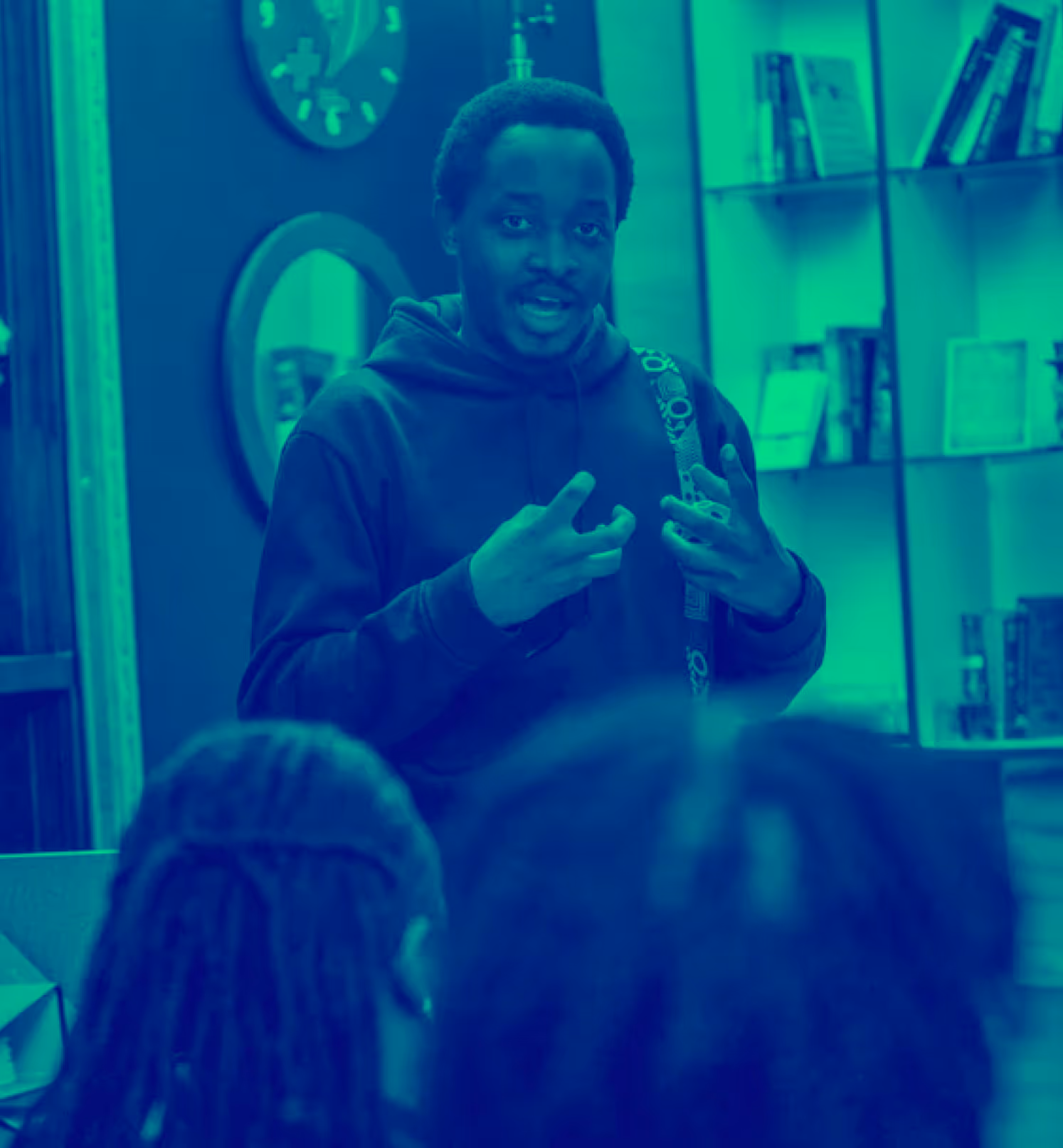 A man in a hoodie speaking and gesturing with hands to an audience in a cozy indoor setting.
