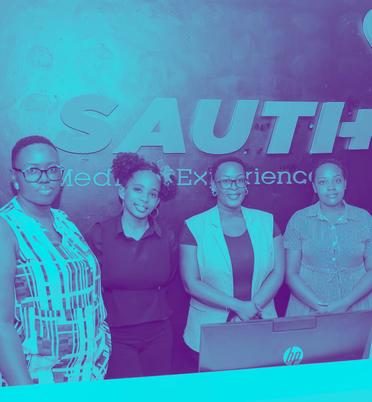 Four women standing behind a desk in front of a wall with large text reading 'SAUTI+'