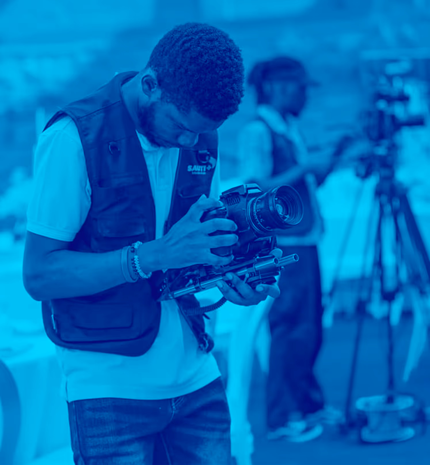 Man in vest adjusting professional video camera with another cameraman blurred in background.