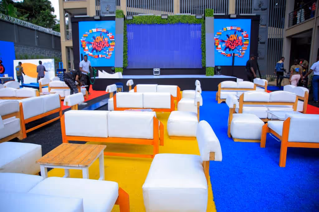 Outdoor event seating with white cushioned chairs and sofas on yellow, blue, and red carpets near stage.