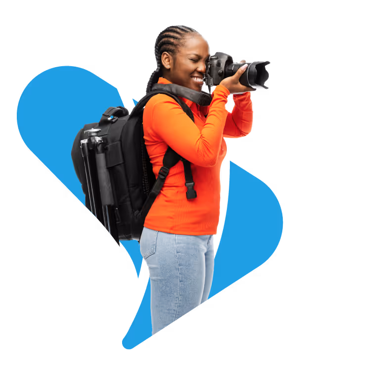 Woman in orange shirt with backpack holding a camera taking a photo, smiling.