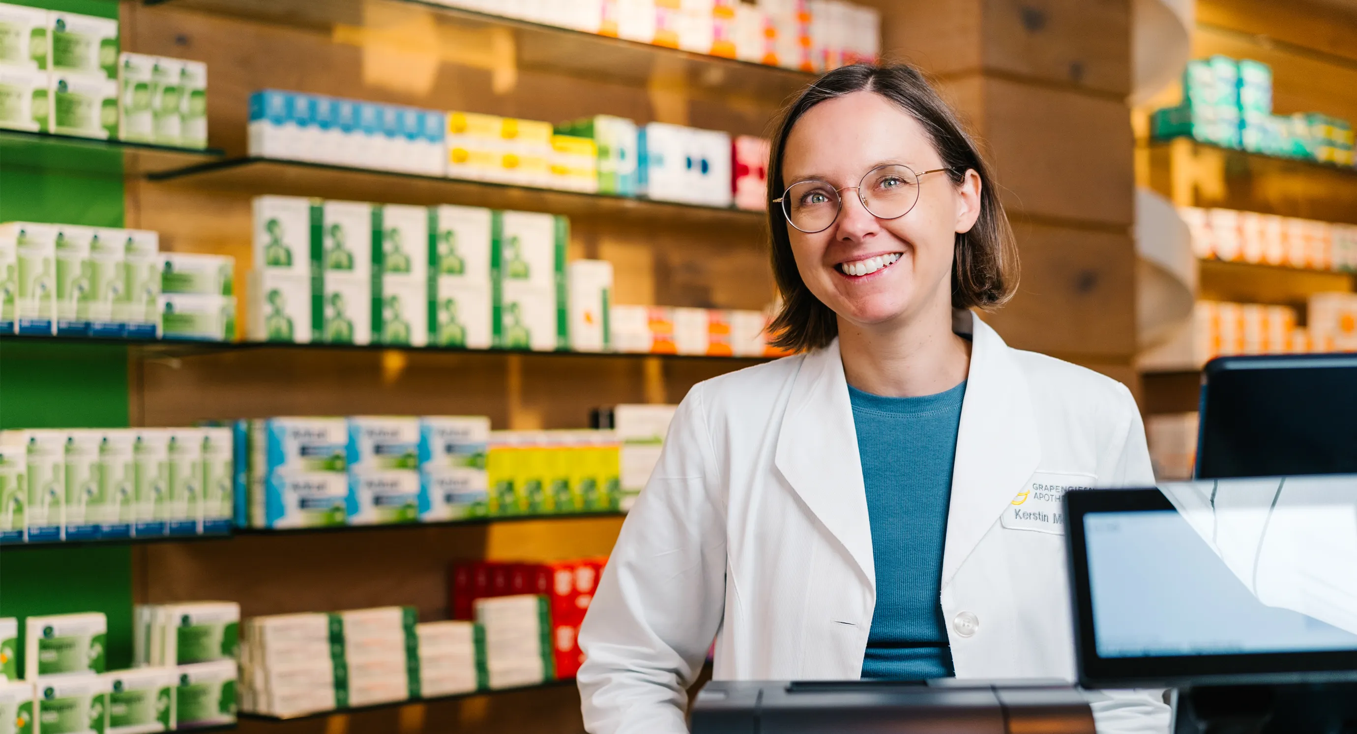 Kerstin Morawe, Pharmazeutisch-technische Assistentin bei der Grapengießer-Apotheke in Lüneburg, steht stellvertrendend für das gesamte Team lächelnd hinter dem HV-Tisch der modernen Apotheke. Im Hintergrund sind Regale mit Arzneimitteln und Gesundheitsprodukten zu sehen – ein Ort, an dem persönliche Beratung, pharmazeutische Kompetenz und digitale Services wie E-Rezept und Botendienst im Mittelpunkt stehen.